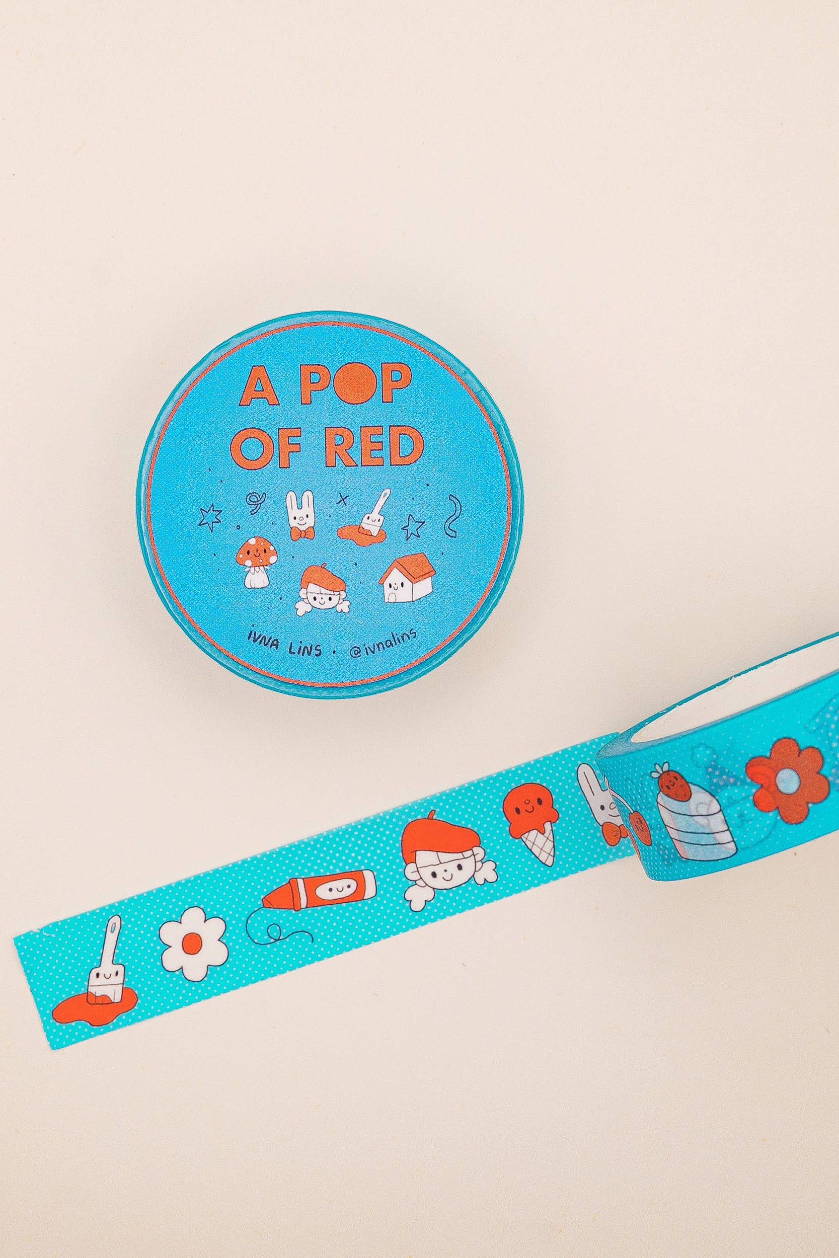 Blue washi tape with cartoon designs and a round container labeled 'A Pop of Red' on a beige background