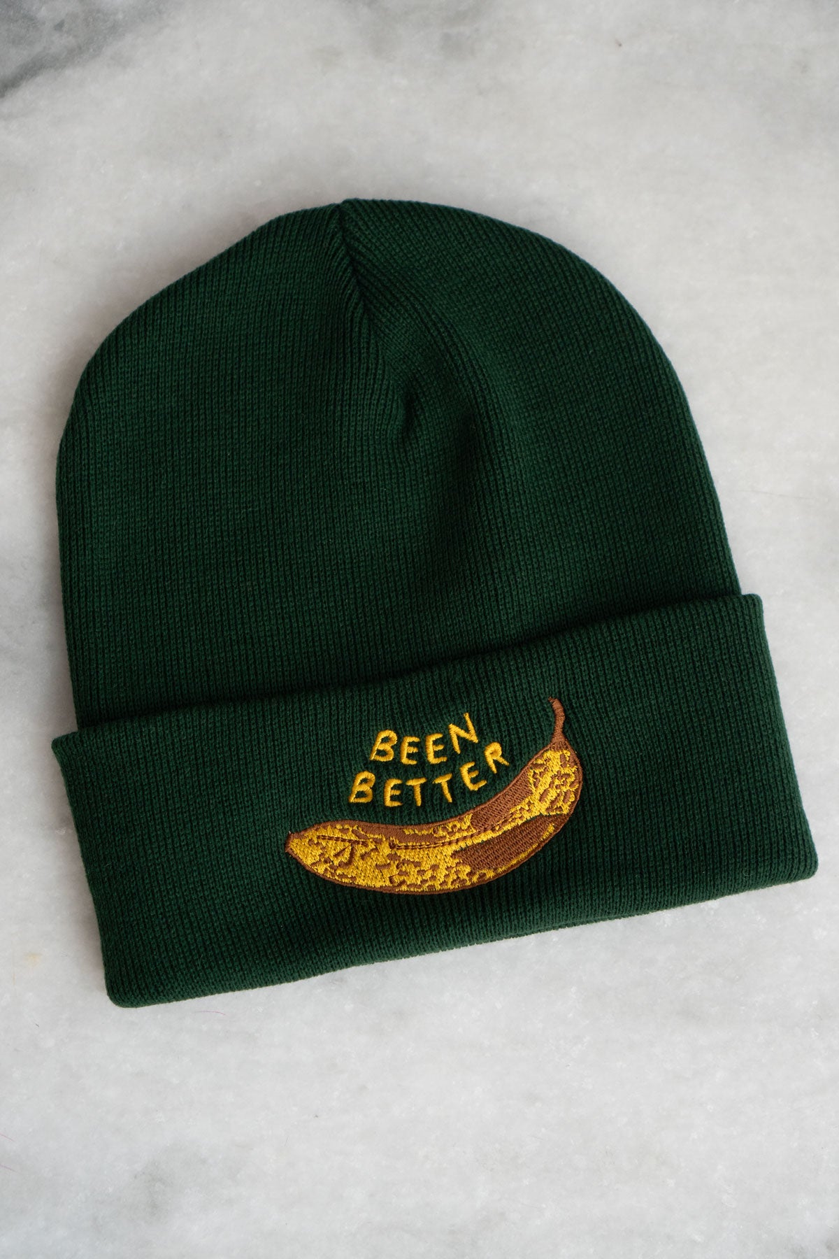 Forest green knit beanie with embroidery of a bruised banana and the phrase "been better" on the rolled cuff