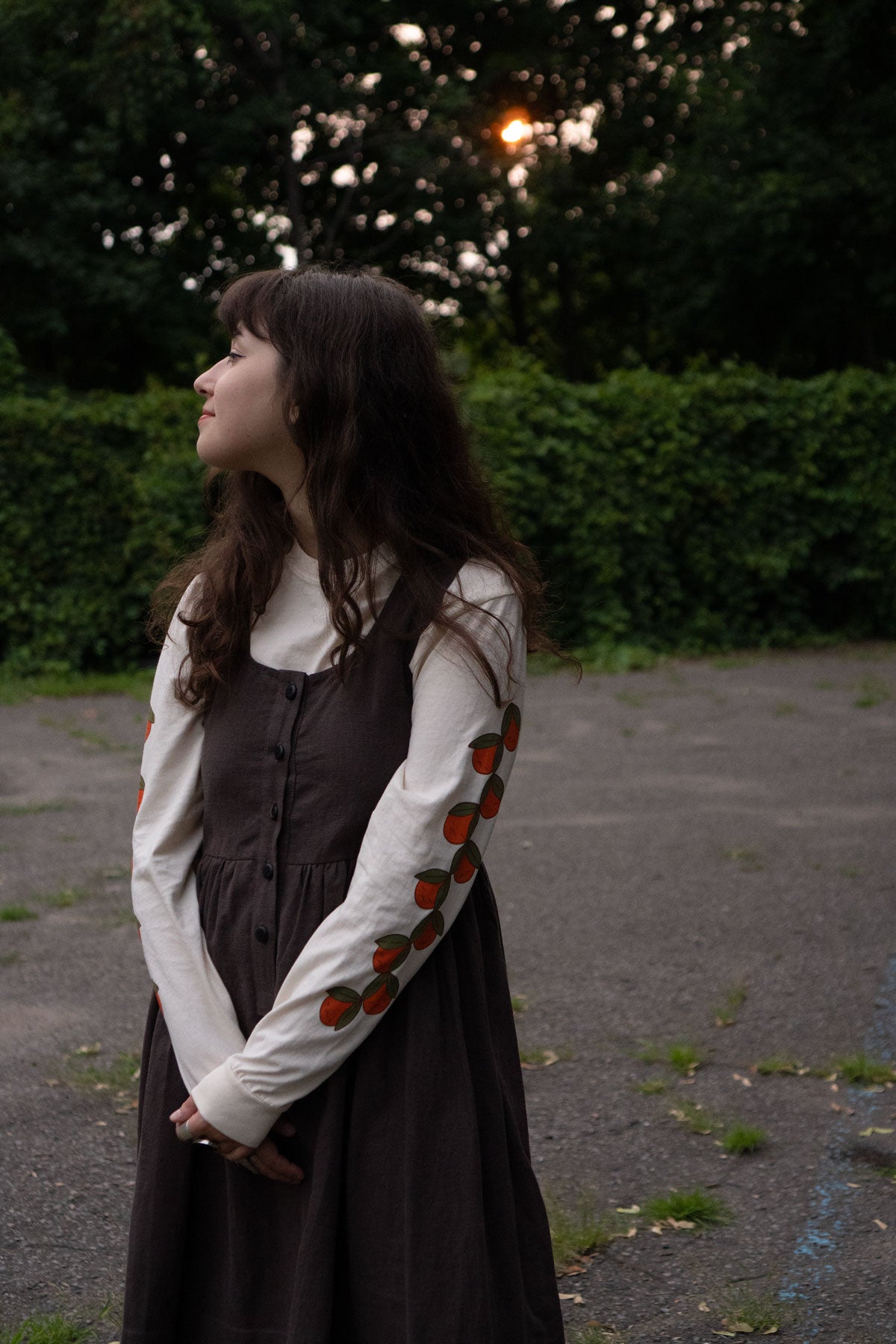 Woman in a brown dress layerd over an off-white long sleeve t-shirt featuring fruit patterns standing outdoors.