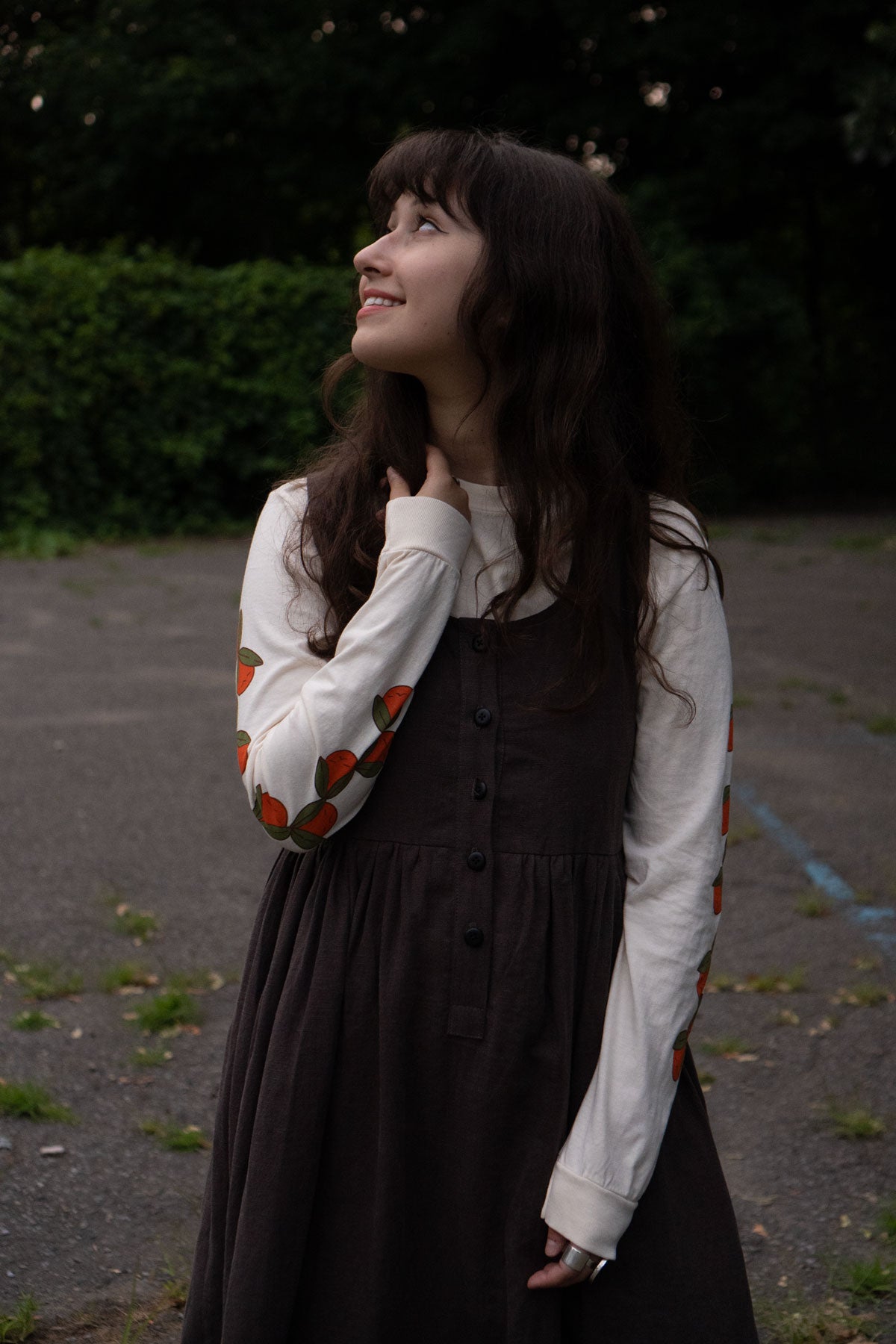 Woman in a brown dress and off-white long sleeved shirt with orange patterns standing outdoors.