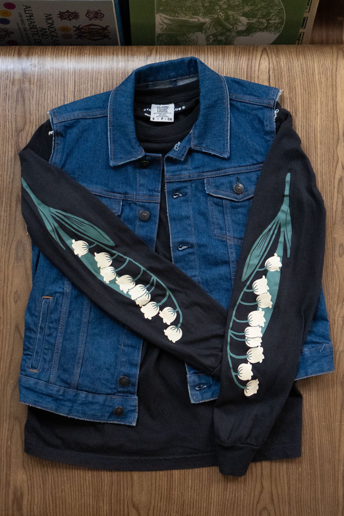 Black long-sleeved tee with lily of the valley flowers prited down the sleeves layered under a denim vest
