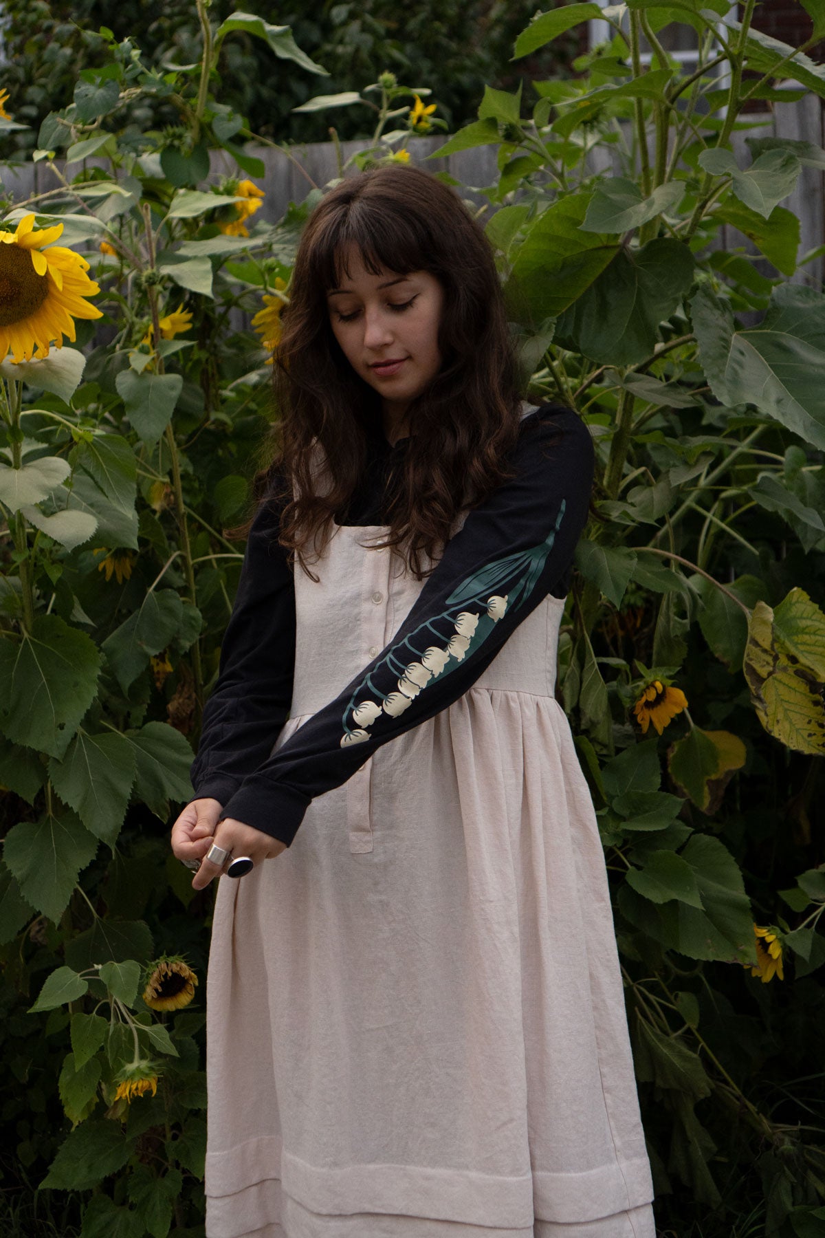 Woman in a garden with sunflowers wearing a dress and blacklong-sleeved t-shirt with floral print sleeves.