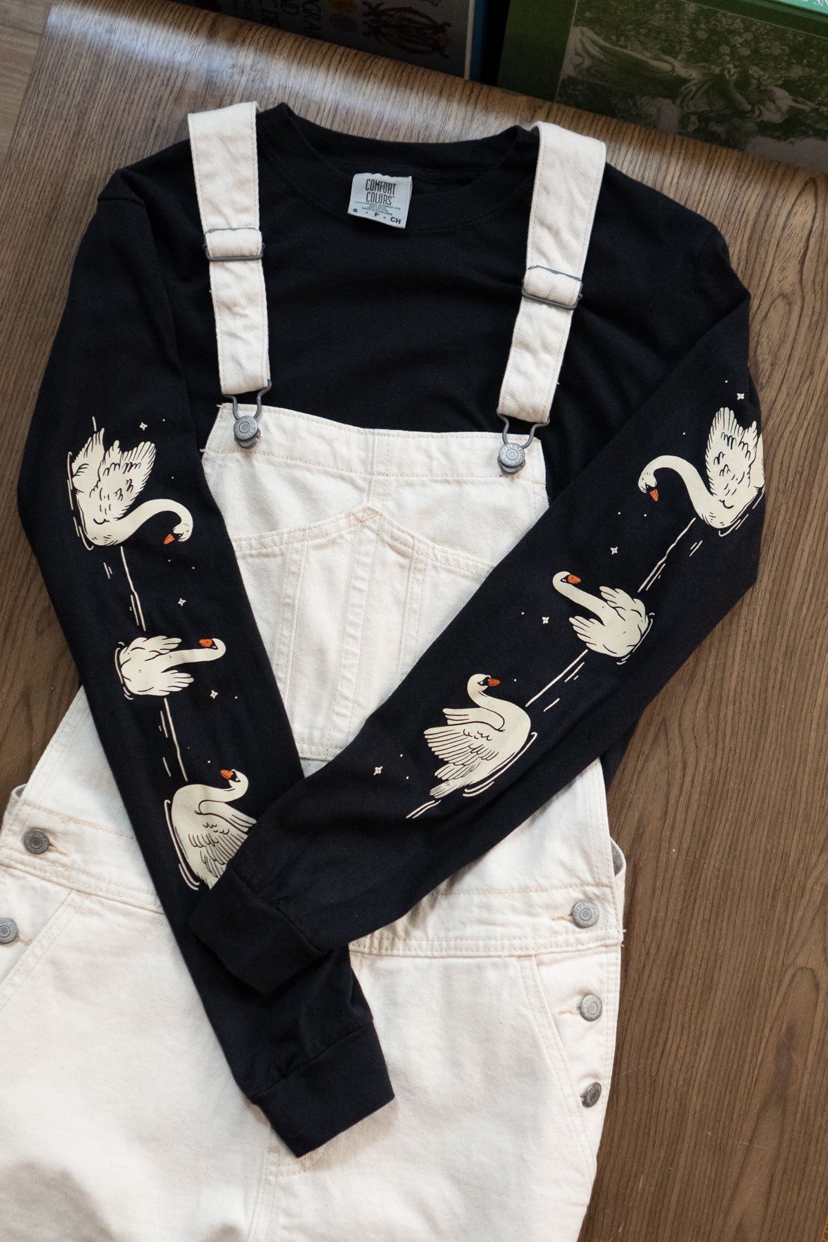 Black long-sleeved t-shirt with swans printed down each arm layered under off-white overalls.