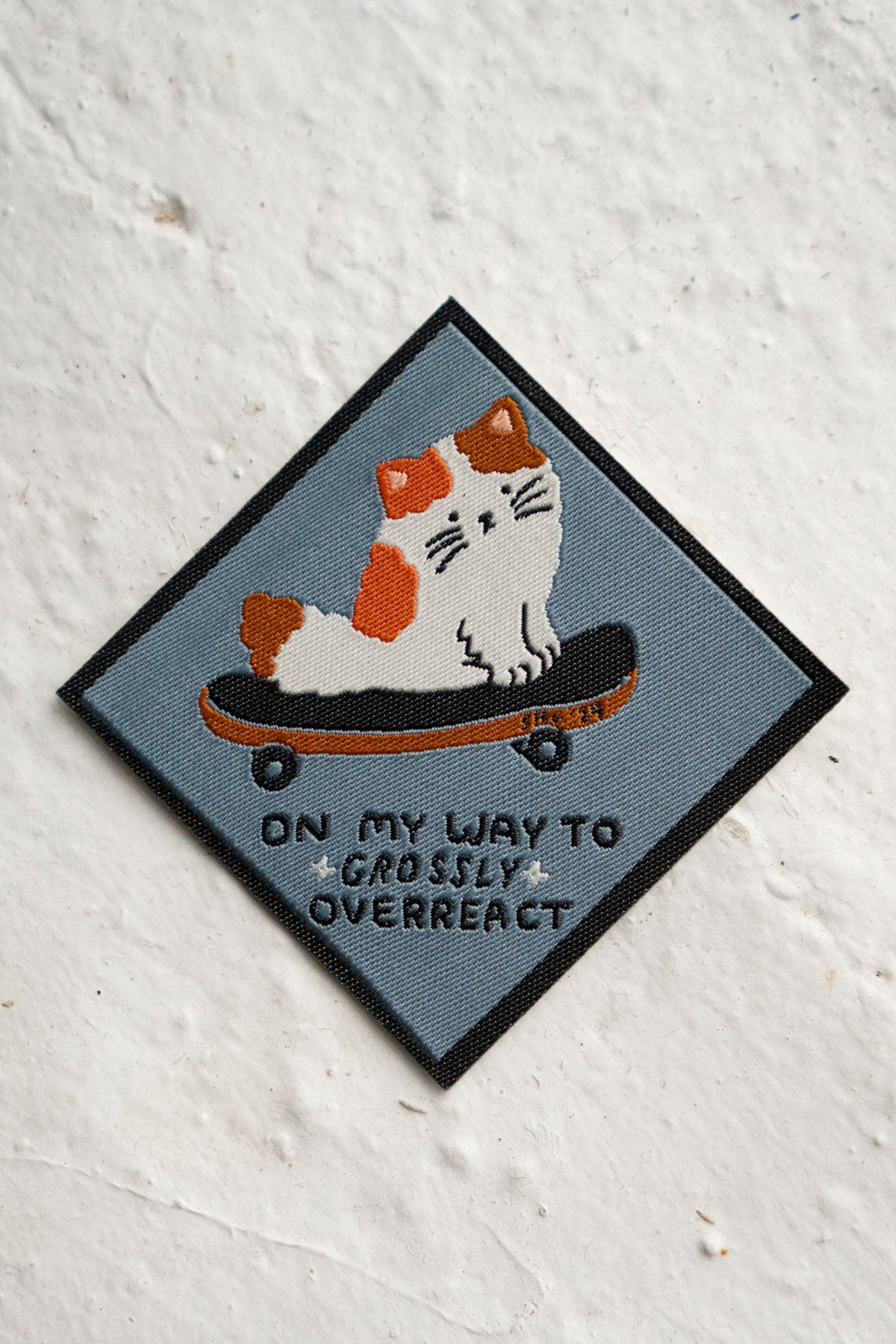 Woven patch in diamond shape featuring a cute fluffy cat on a skateboard and the phrase "On my way to grossly overreact"