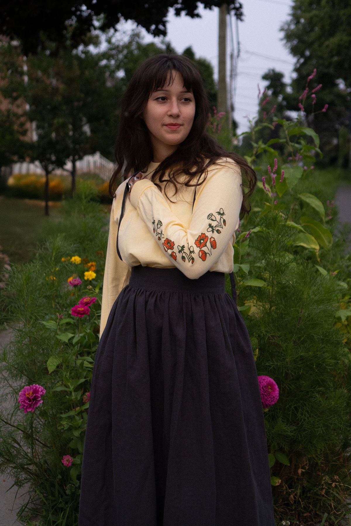Woman wearing a long-sleeved shirt with floral print sleeves and dark skirt standing in a garden.