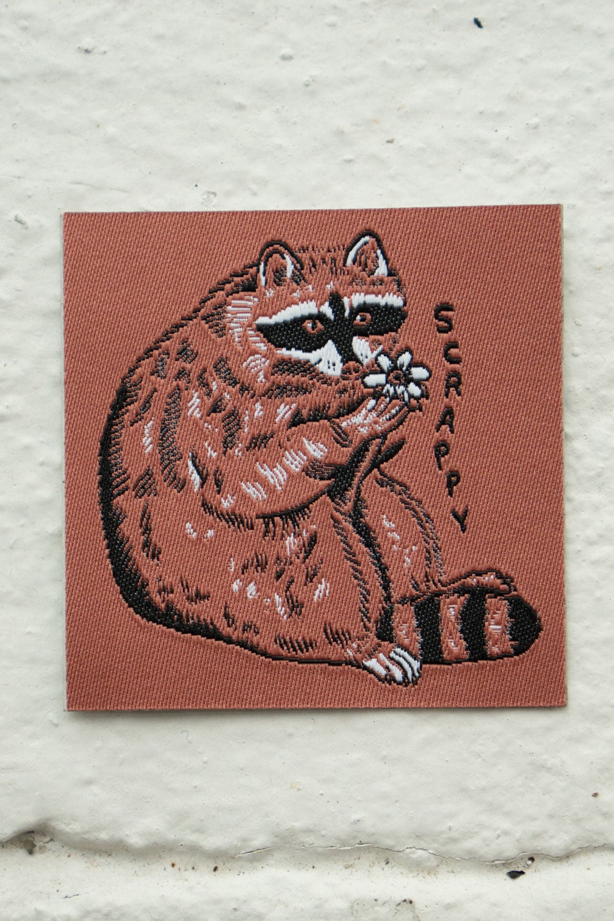 Woven patch in a copper tone with an image of a raccoon holding a flower and text saying "Scrappy"