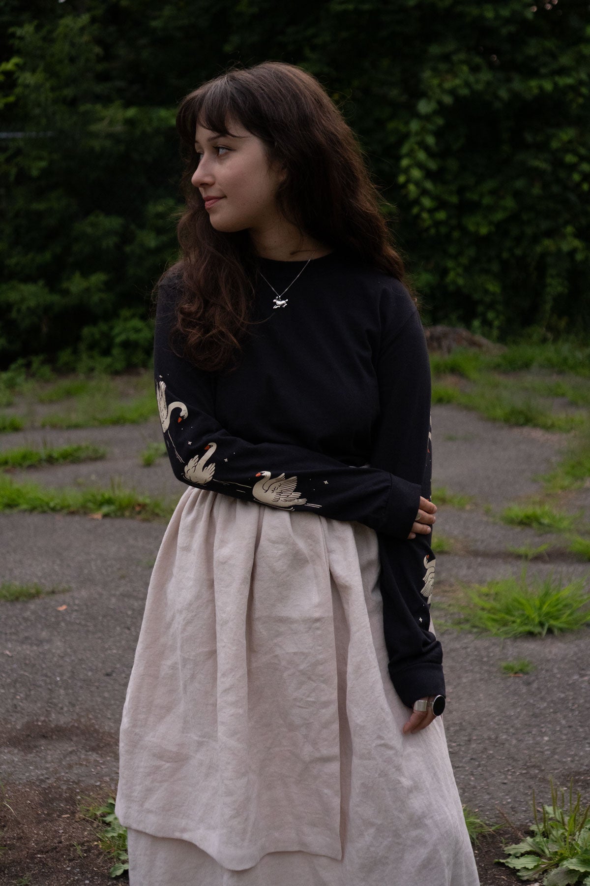 Woman wearing a black long-sleeved t-shirt with swans printed on the sleeves and beige skirt standing outdoors with greenery in the background