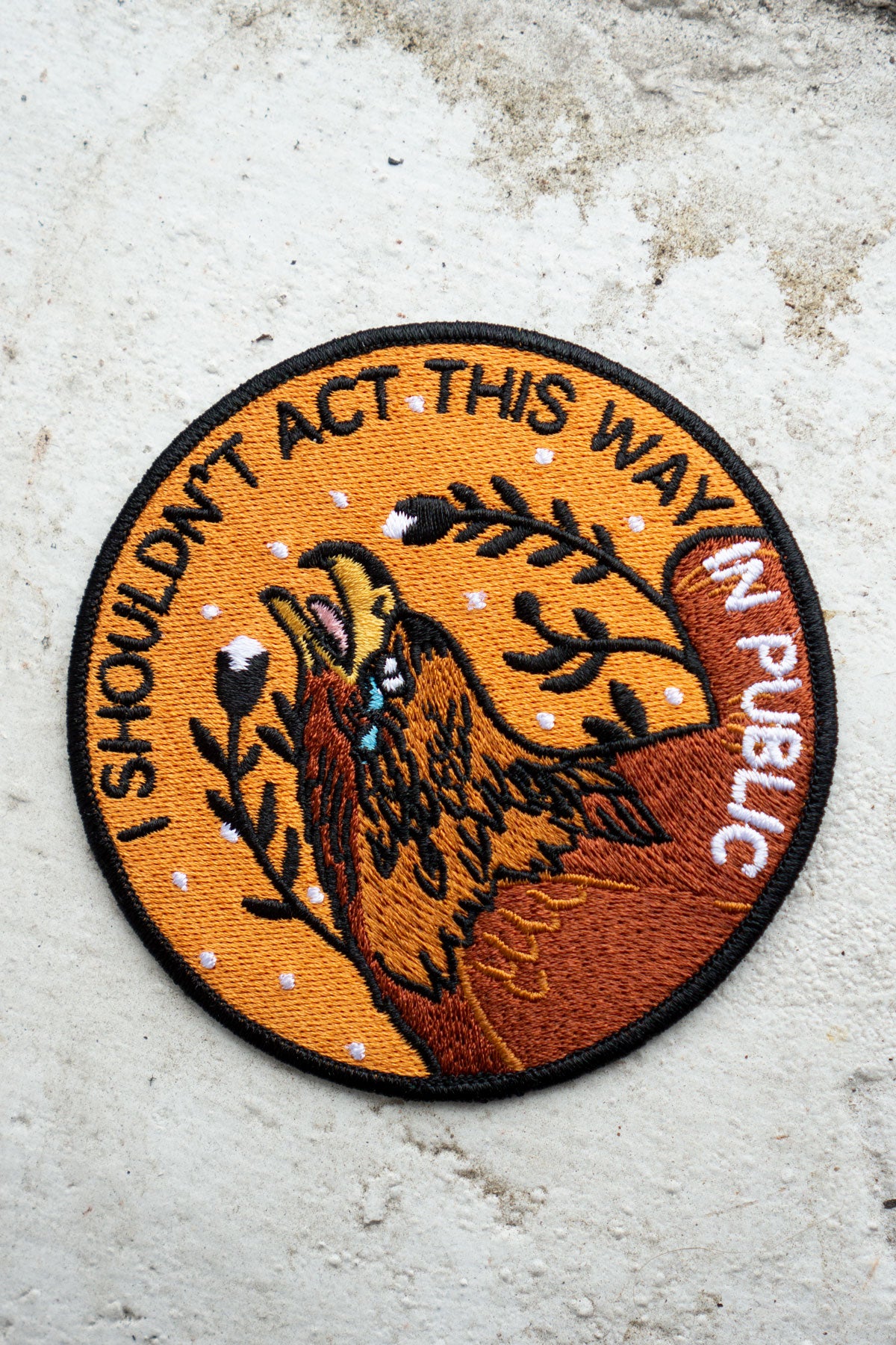 Round embroidered patch in tones of yellow and brown with imagery of a crying eagle and the words "I Shouldn't Act This Way in Public"